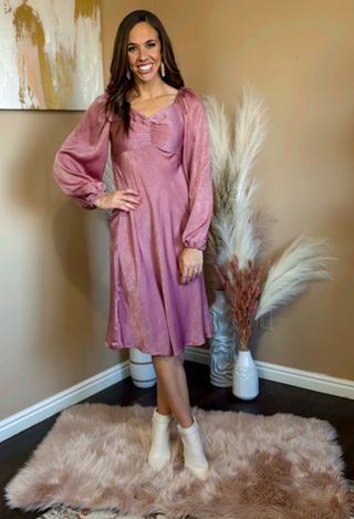 A woman standing and posing in a pink knee-length dress with a sweetheart neckline and long sleeves, on a faux fur rug.